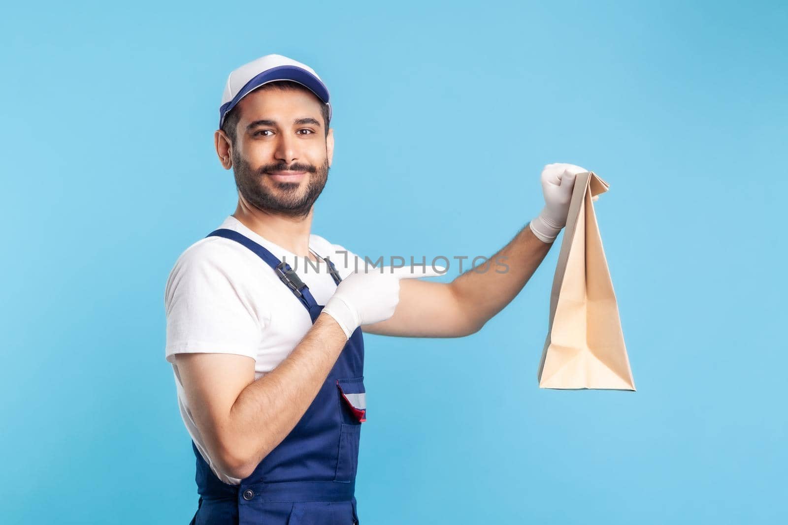 Look at bag. Portrait of cheerful handyman in overalls and gloves pointing at ordered parcel. Courier delivering food in paper package, post mail services. studio shot isolated on blue background