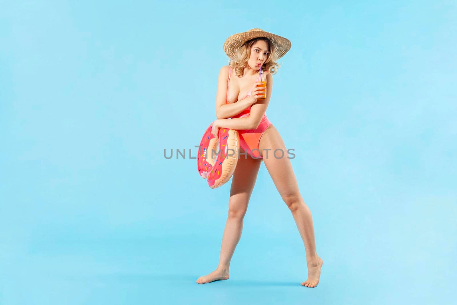 Full length sexy beautiful woman in swimsuit and sun hat holding rubber ring, drinking fresh juice or cocktail, enjoying rest on summer beach, vacation resort. studio shot isolated on blue background