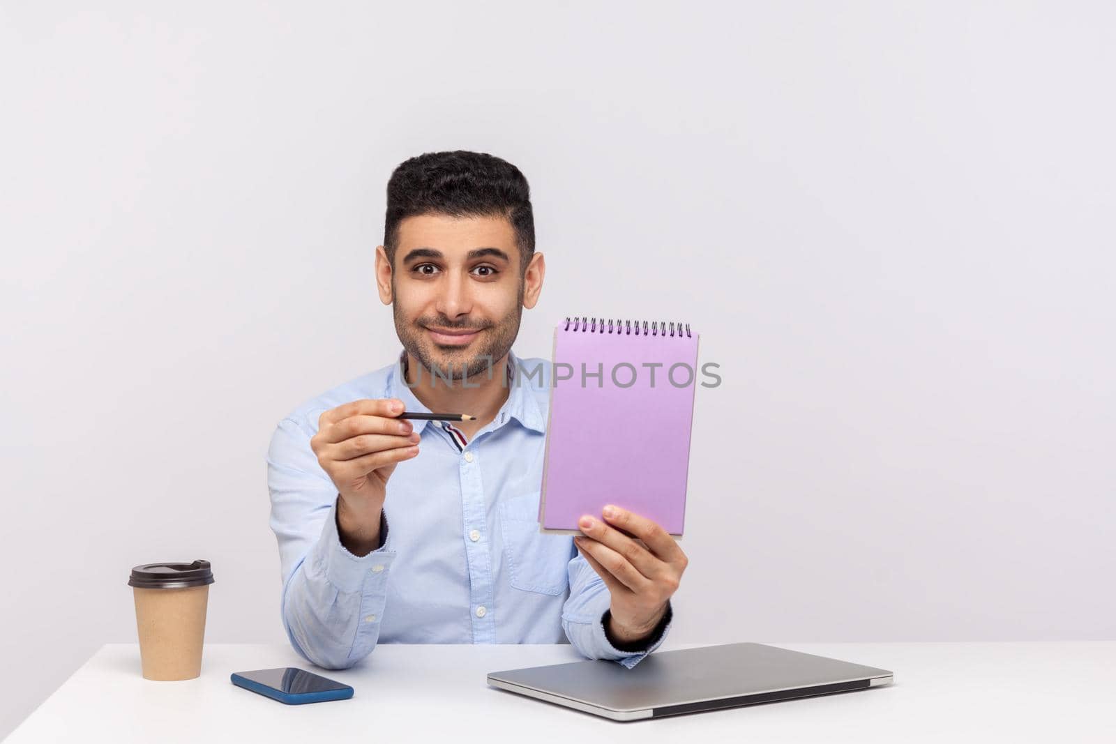 Happy businessman sitting in office workplace, pointing empty sheet and smiling at camera, showing paper notebook template, mock up blank space for business idea, message. indoor studio shot isolated
