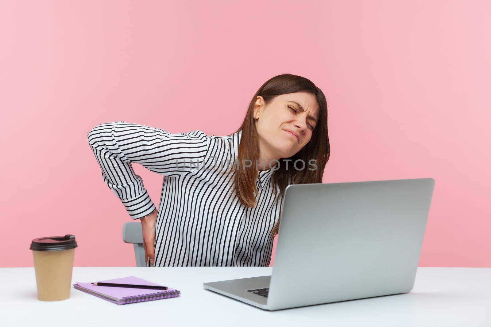 Unhappy dissatisfied woman office worker touching and massaging spine sitting at workplace, feeling ache in lower back, has problems with kidneys. Indoor studio shot isolated on pink background