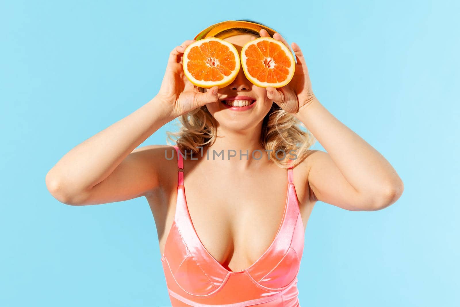 Portrait of cheerful smiling woman in swimsuit covering eyes with fresh cut orange, having fun. Concept of female beauty and skin care, natural cosmetic antioxidant. indoor studio shot isolated