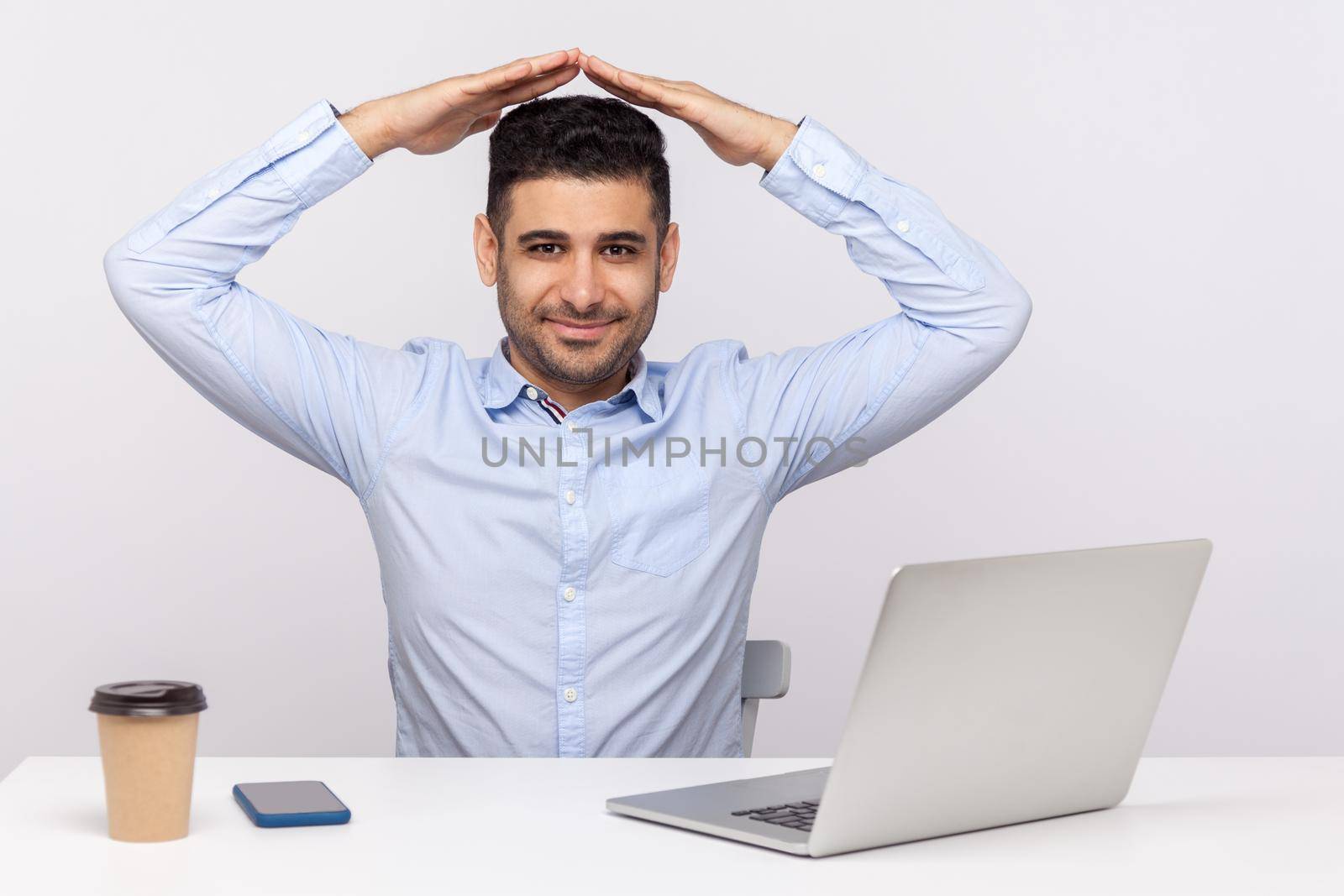 Happy elegant man employee, insurance agent sitting in office workplace with laptop, keeping house roof gesture over head and smiling at camera. indoor studio shot isolated on white background