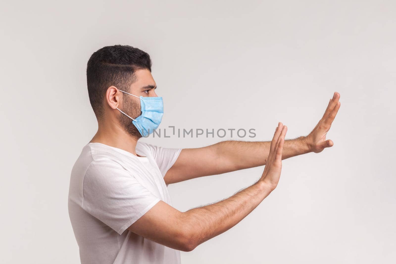 Side view of alarming man in hygienic protective mask gesturing stop, afraid of coronavirus infection, respiratory illnesses such as flu, 2019-nCoV. indoor studio shot isolated on white background