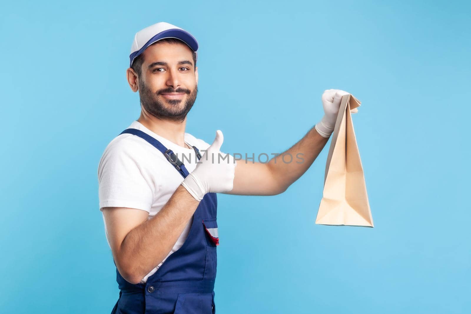 Excellent job. Portrait of satisfied handyman in overalls and gloves holding ordered parcel, showing thumbs up, recommending delivery, post mail service. indoor studio shot isolated on blue background