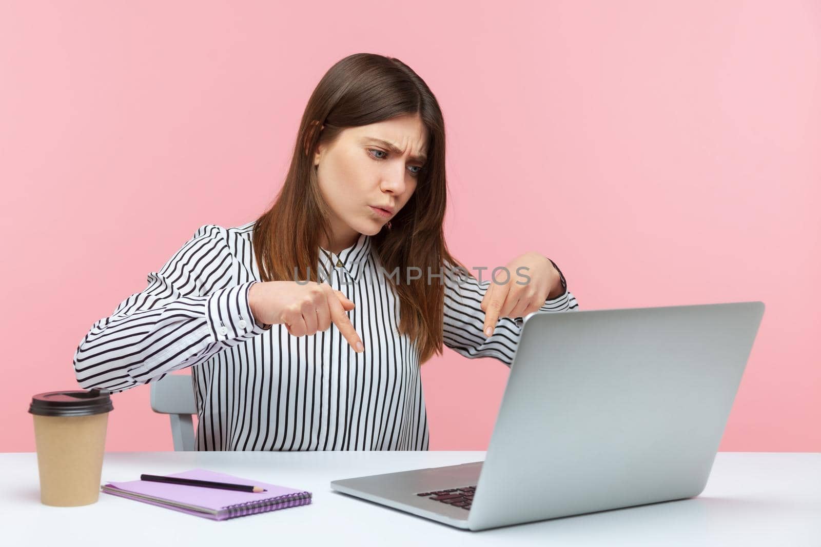 Here and right now. Serious bossy business woman pointing fingers down talking on video call on laptop, controlling and demanding deadline job finish. Indoor studio shot isolated on pink background