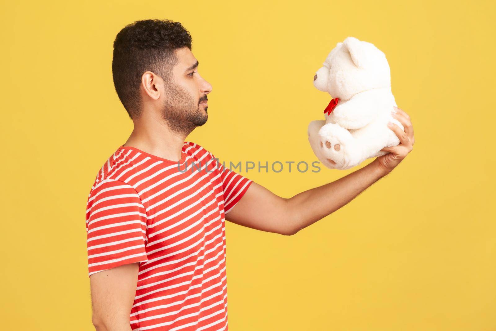 Profile portrait bearded man holding in hands and looking at white toy bear, enjoying gift and missing his beloved woman. Indoor studio shot isolated on yellow background