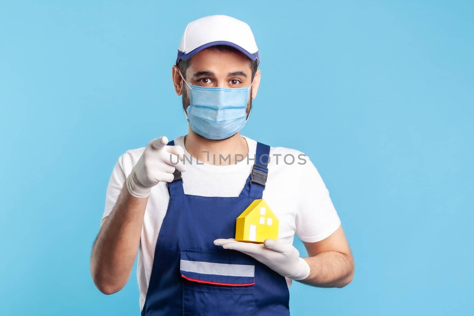 Hey you. Portrait of happy handyman, mover guy in workwear, mask and gloves holding paper house and pointing to camera. Advertising home repair, maintenance or moving services, help in relocation.