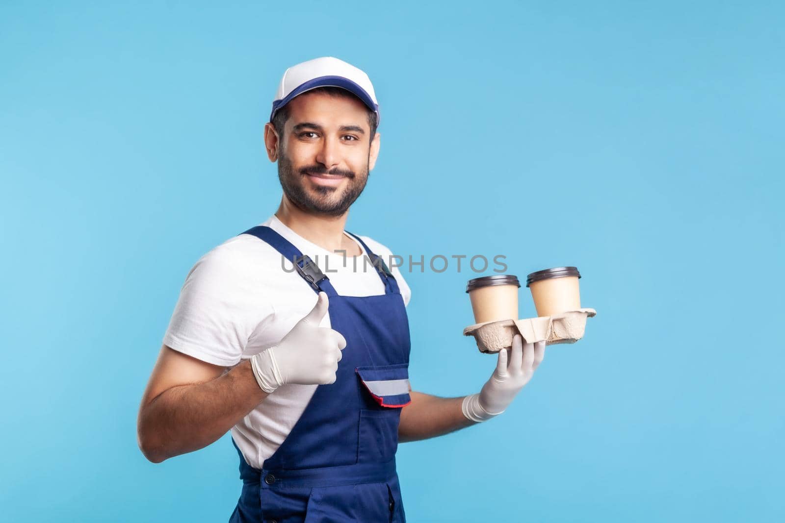 Delivery service. Side view, friendly courier in overalls holding coffee, wearing safety gloves offering drinks in disposable cups and showing thumbs up, like gesture. indoor studio shot, isolated
