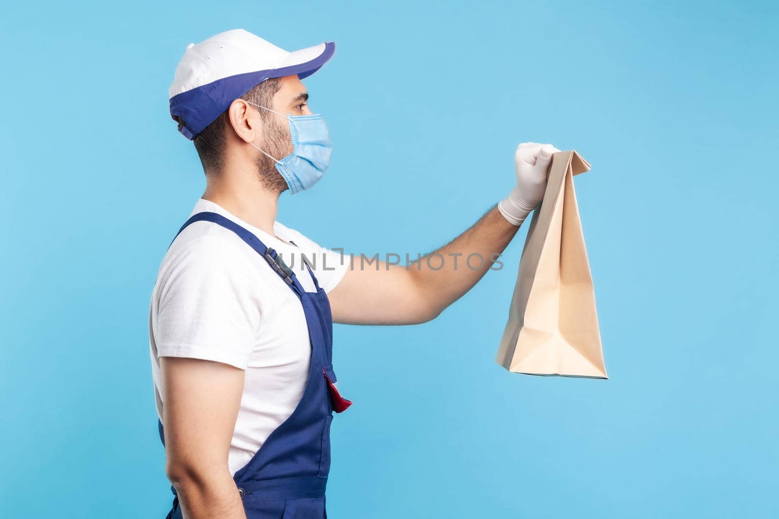 Side view, friendly professional courier in uniform, mask and safety gloves giving groceries bag, delivering takeaway food, post services express shipping. indoor shot isolated on blue background