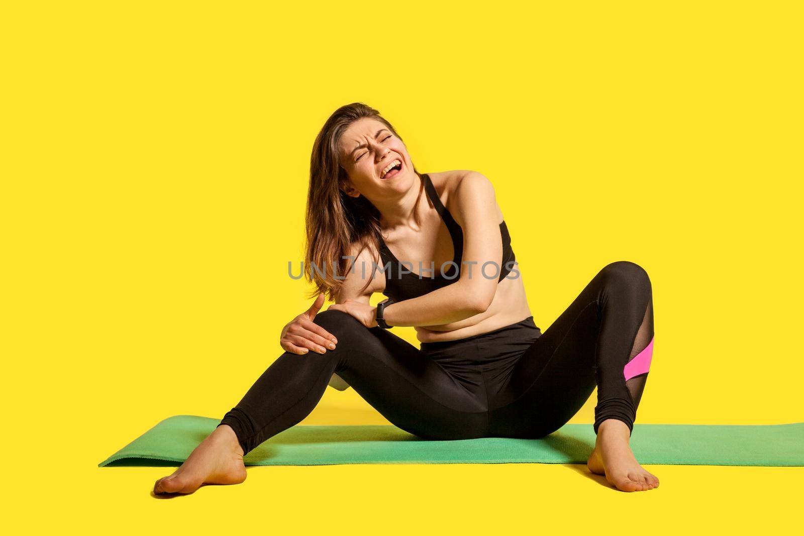 Sportswoman shouting from knee pain, sitting on gym mat, touching leg suffering joint injury after workout, muscle strain, hurting to move sprained ligaments. studio shot isolated on yellow background