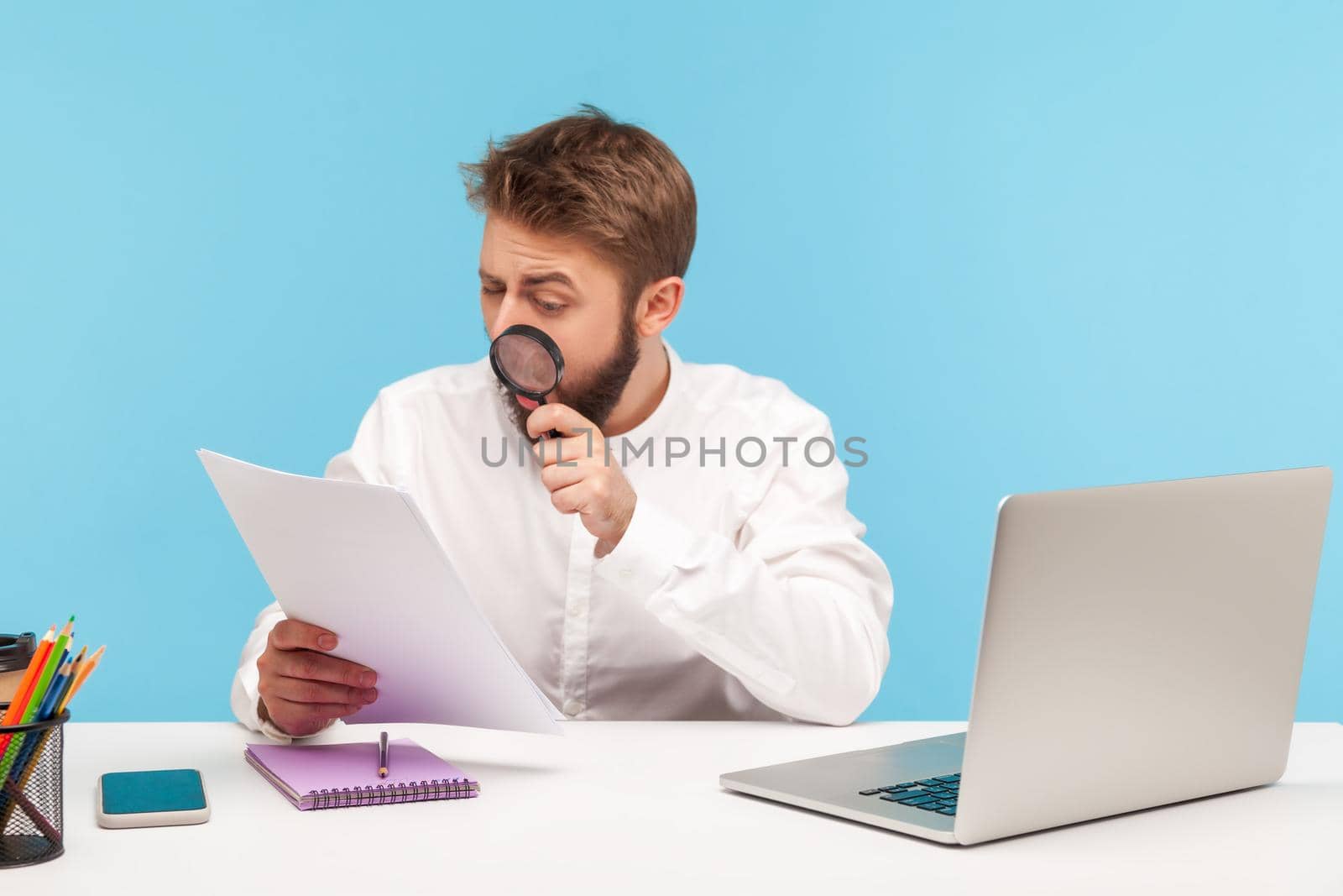 Serious businessman attentively studying analyzing papers holding magnifying glass, looking for mistakes and errors, checking datas sitting at workplace. Indoor studio shot isolated on blue background