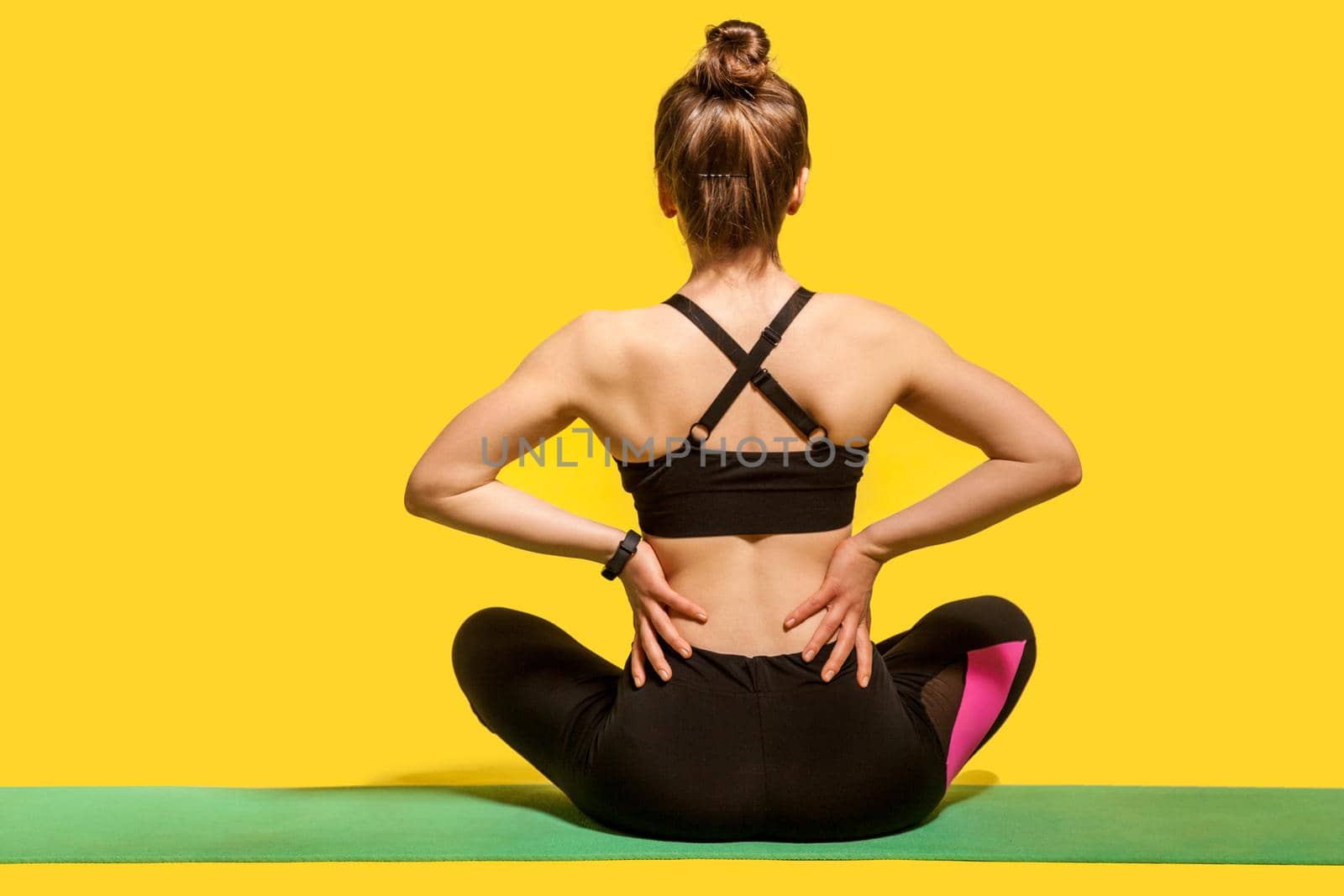 Back view, sportswoman sitting on mat and touching sore spine, suffering backache after physical training or yoga practice, massaging stiff muscles. indoor studio shot isolated on yellow background