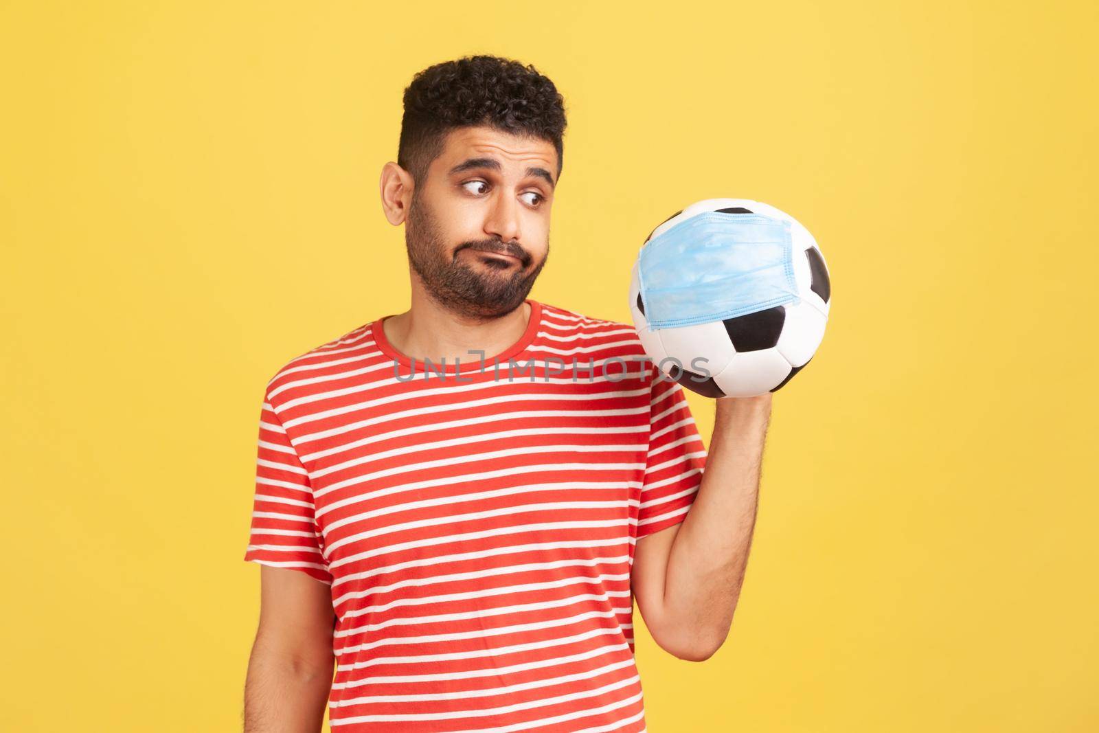 Upset man in red t-shirt looking at soccer ball in safety face mask with disappointment, nervous because of football championship results falsification. Studio shot isolated on yellow background