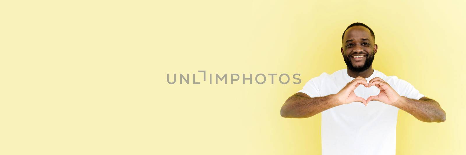 Web banner. Portrait of a handsome cheerful afro american man making a heart shape with his hands, expressing love feelings or friendship, smiling in love on a beige yellow background