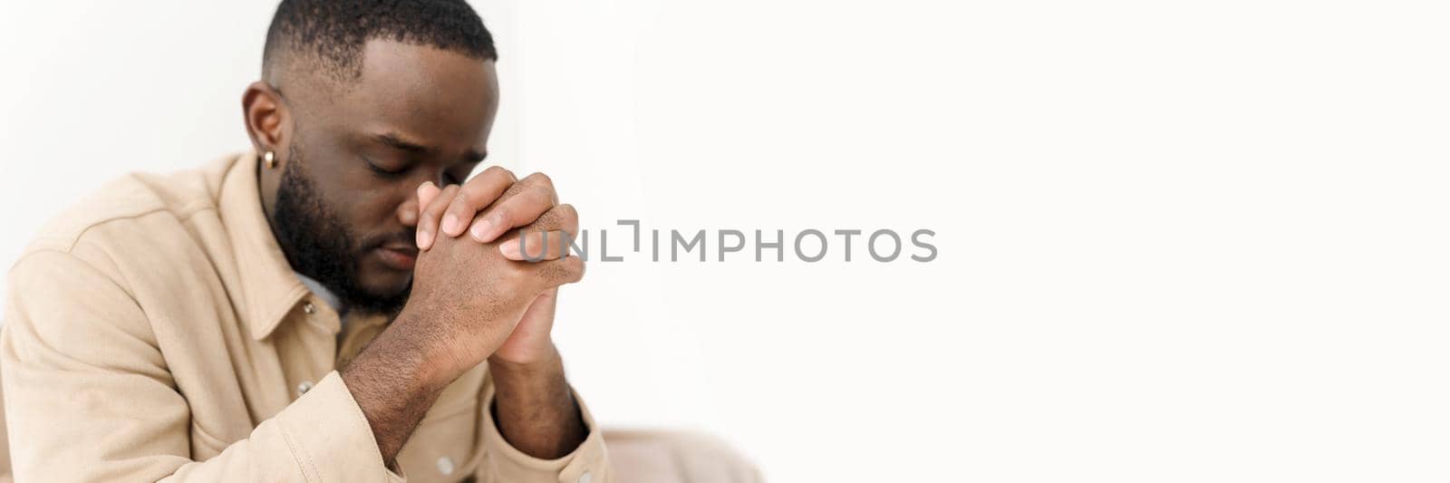 African American man praying for peace while sitting on the couch at home. Web banner