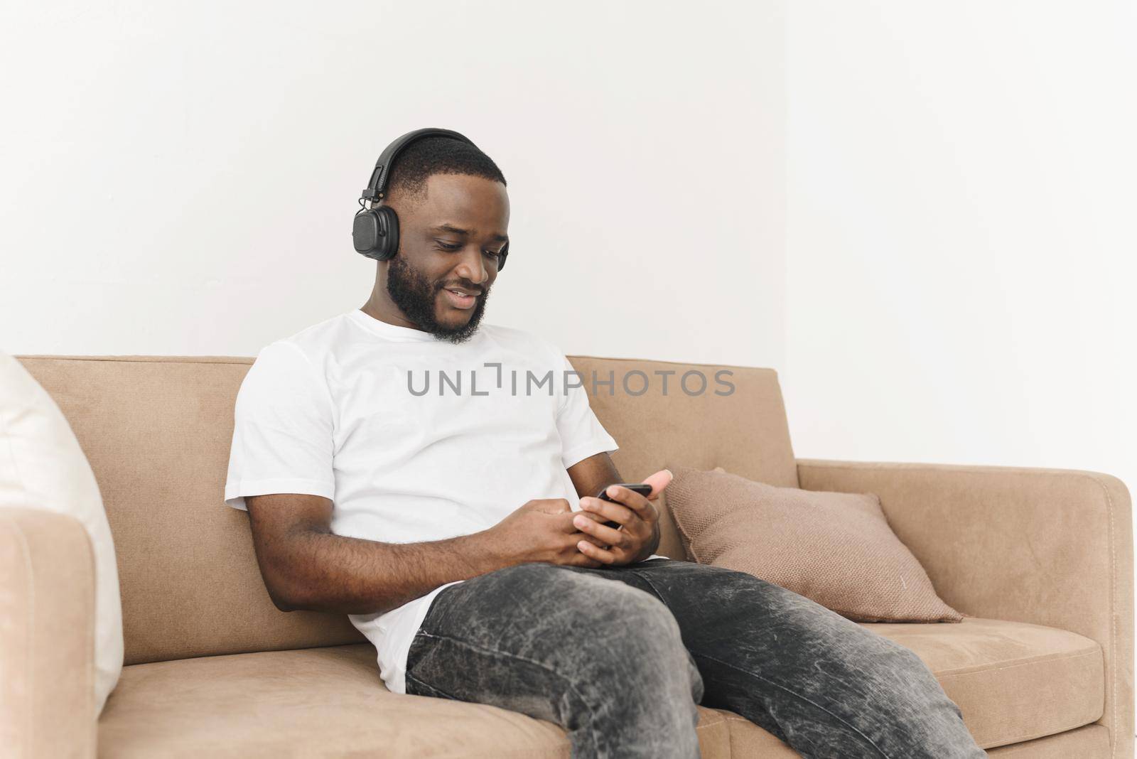 Smiling young african american man in headphones listening to mobile music, browsing social networks using smartphone, relaxing while sitting on sofa at home.