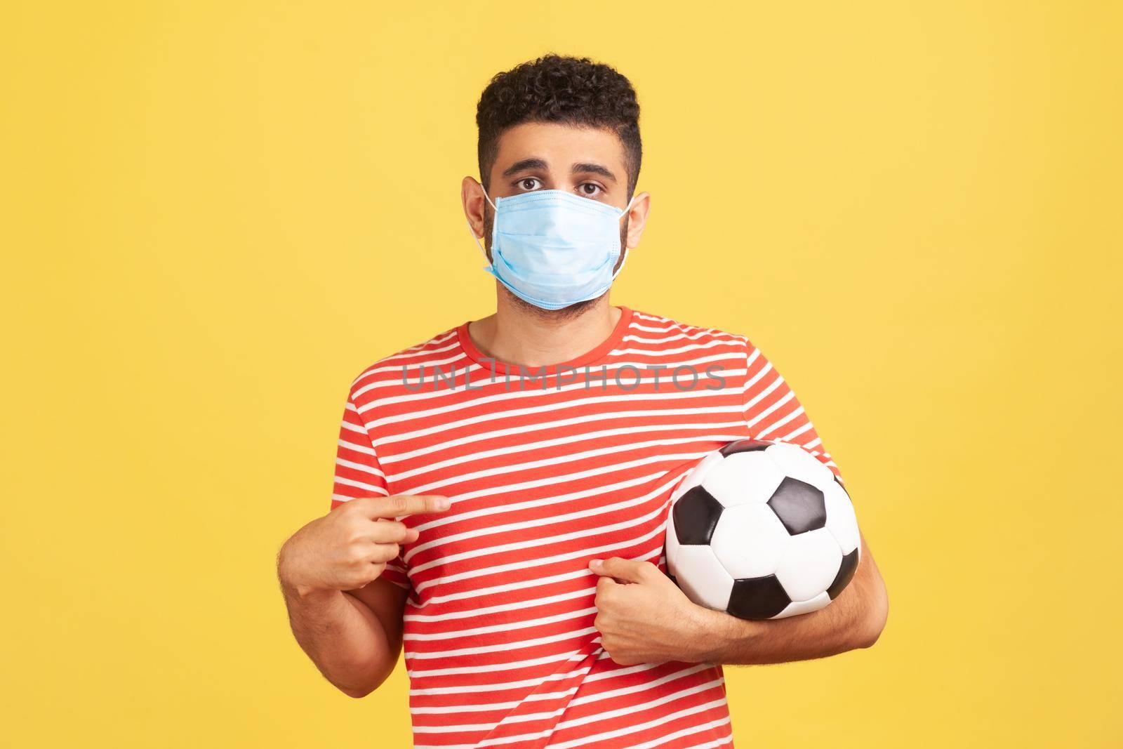 Young bearded man in safety mask and red t-shirt pointing finger at soccer ball on his hand with sad expression, coronavirus quarantine. Indoor studio shot isolated on yellow background