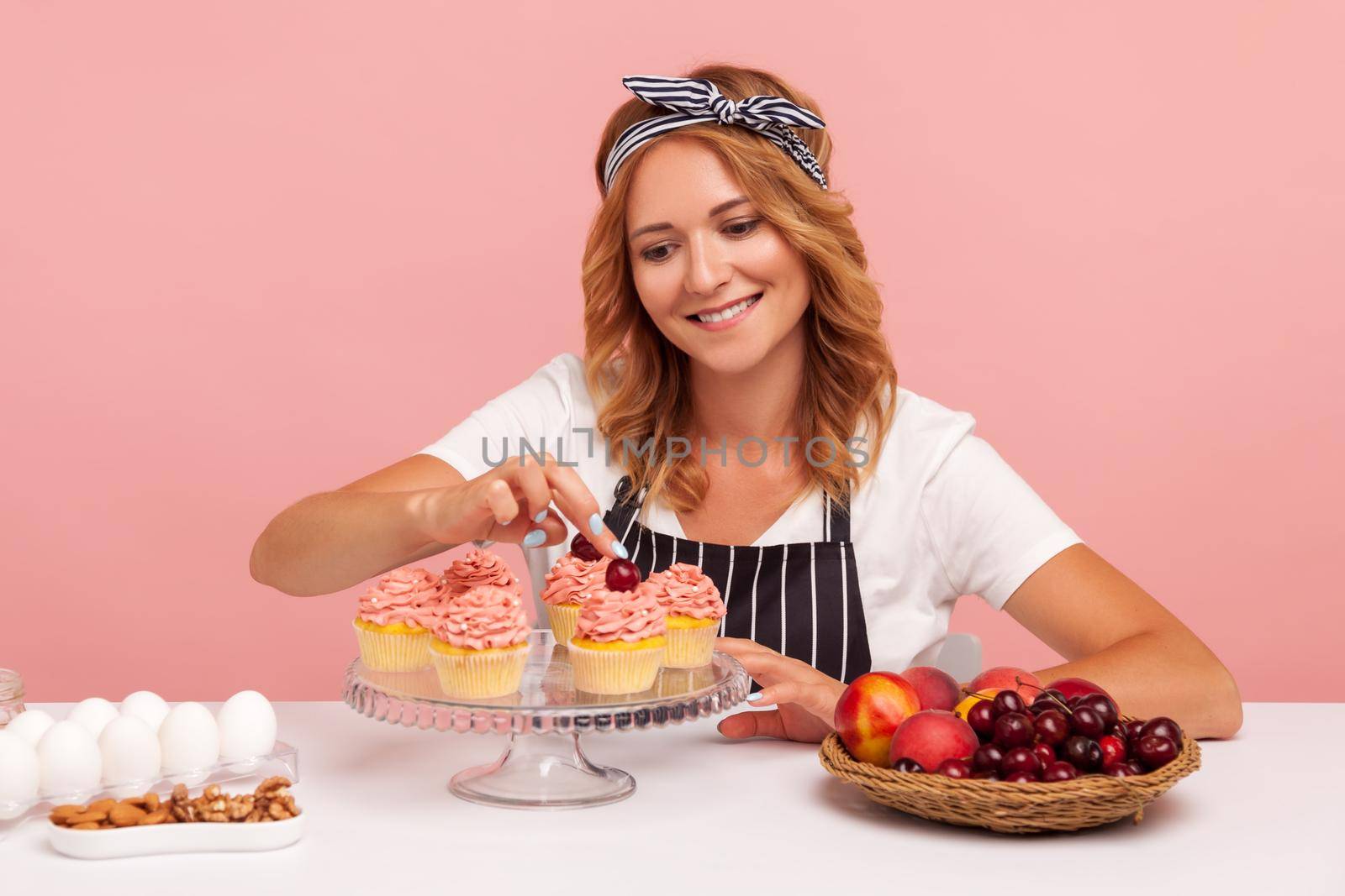 Decoration of finished dessert. Pastry chef sprinkles confectionery homemade pastry with fresh cherry, cooking cakes, wearing T-shirt and apron. Indoor studio shot isolated on pink background.