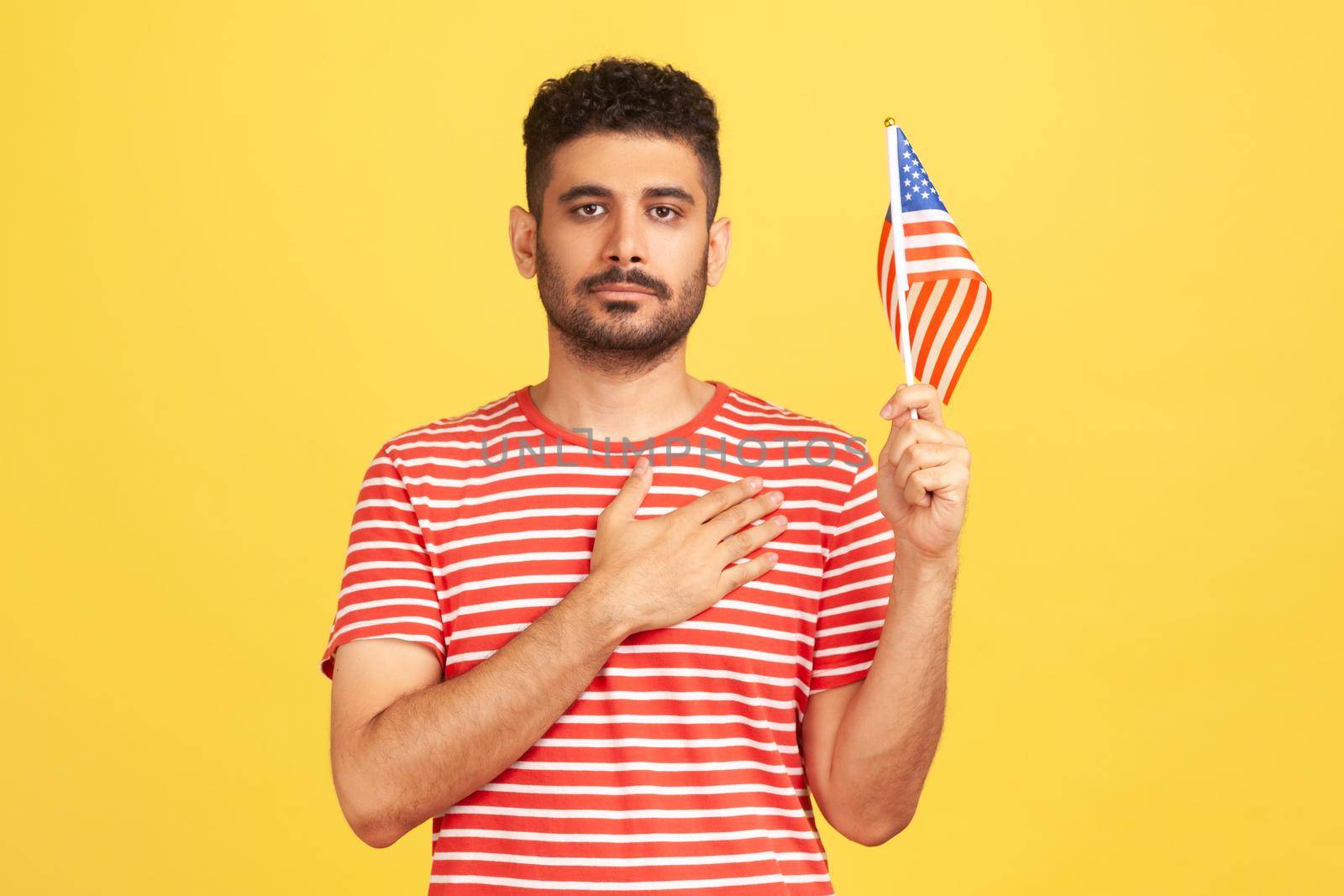 Serious bearded man in striped t-shirt holding united states of america flag holding hand on heart, swearing allegiance, patriotism, independence. Indoor studio shot isolated on yellow background