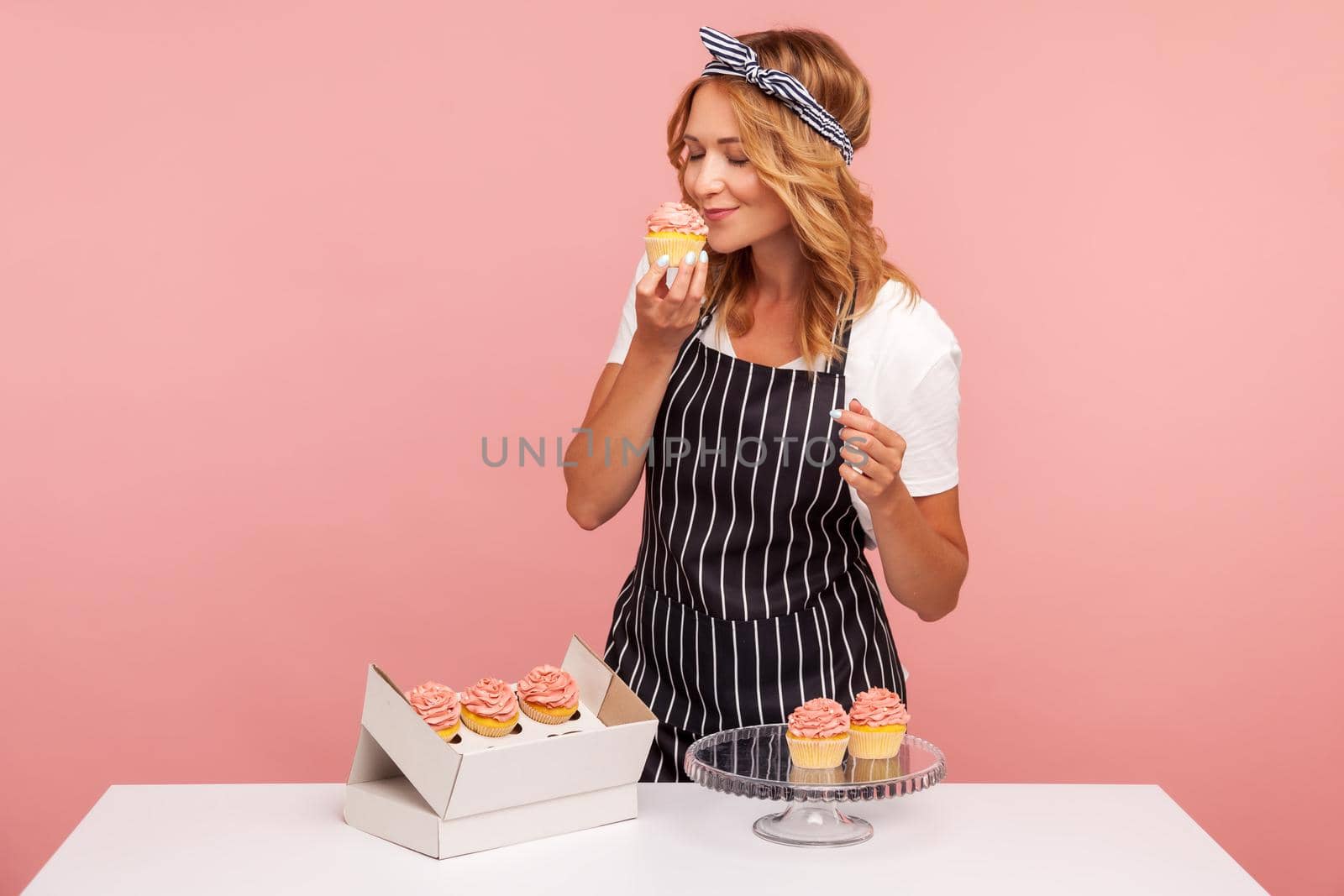 Blonde young adult baker takes out cakes from paper box, preparing for serving to guests, enjoying of smelling tasty dessert with closed eyes. Indoor studio shot isolated on pink background.