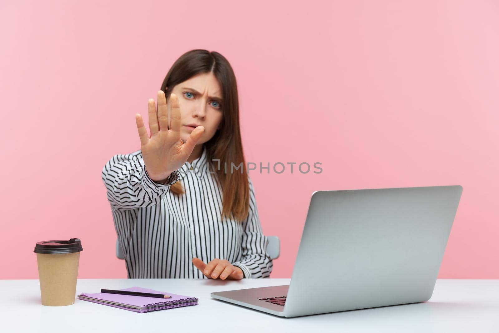 Serious confident business woman in striped shirt holding stop gesture with hand sitting in office, rejecting sexual harassmet at work. Indoor studio shot isolated on pink background