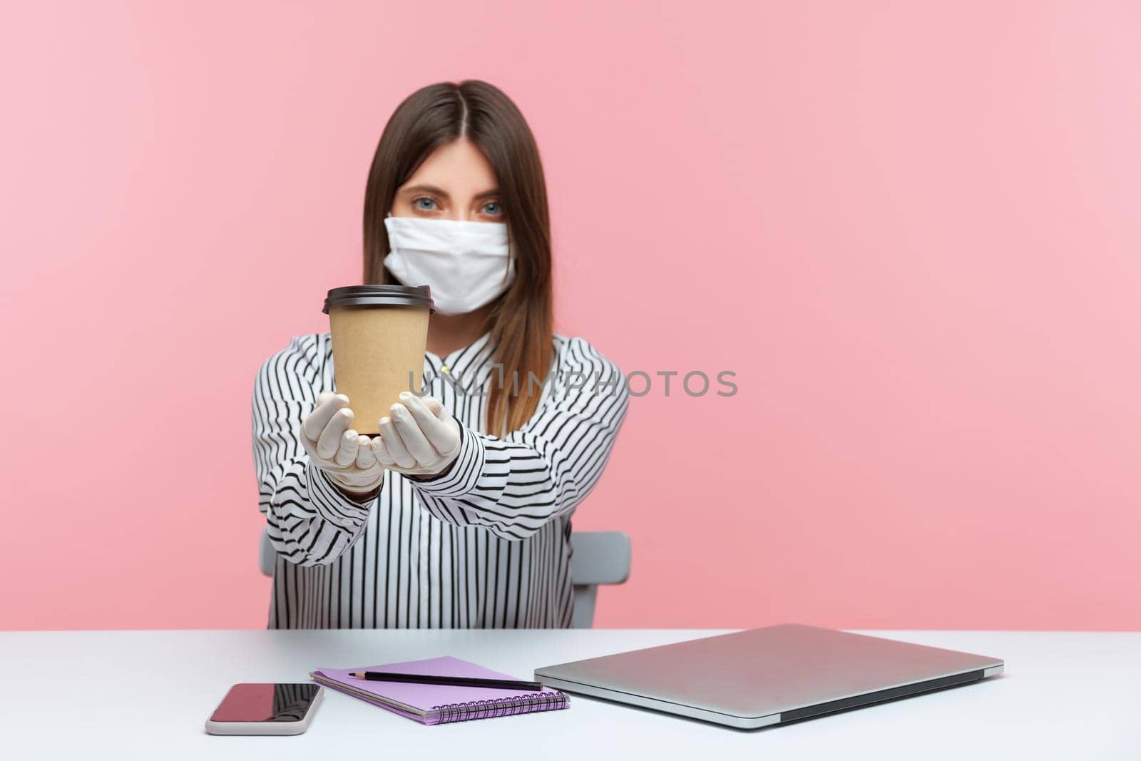 Woman office employee showing cup of coffee, sitting at workplace, feeling safe healthy with hygienic face mask and protective gloves, working in self-isolation, coronavirus quarantine. studio shot