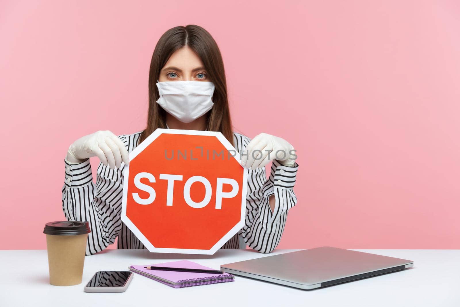 Prohibitions and restrictions during quarantine. Woman office employee sitting safe healthy with hygienic face mask and protective gloves, holding stop road sign. Warning of coronavirus epidemic