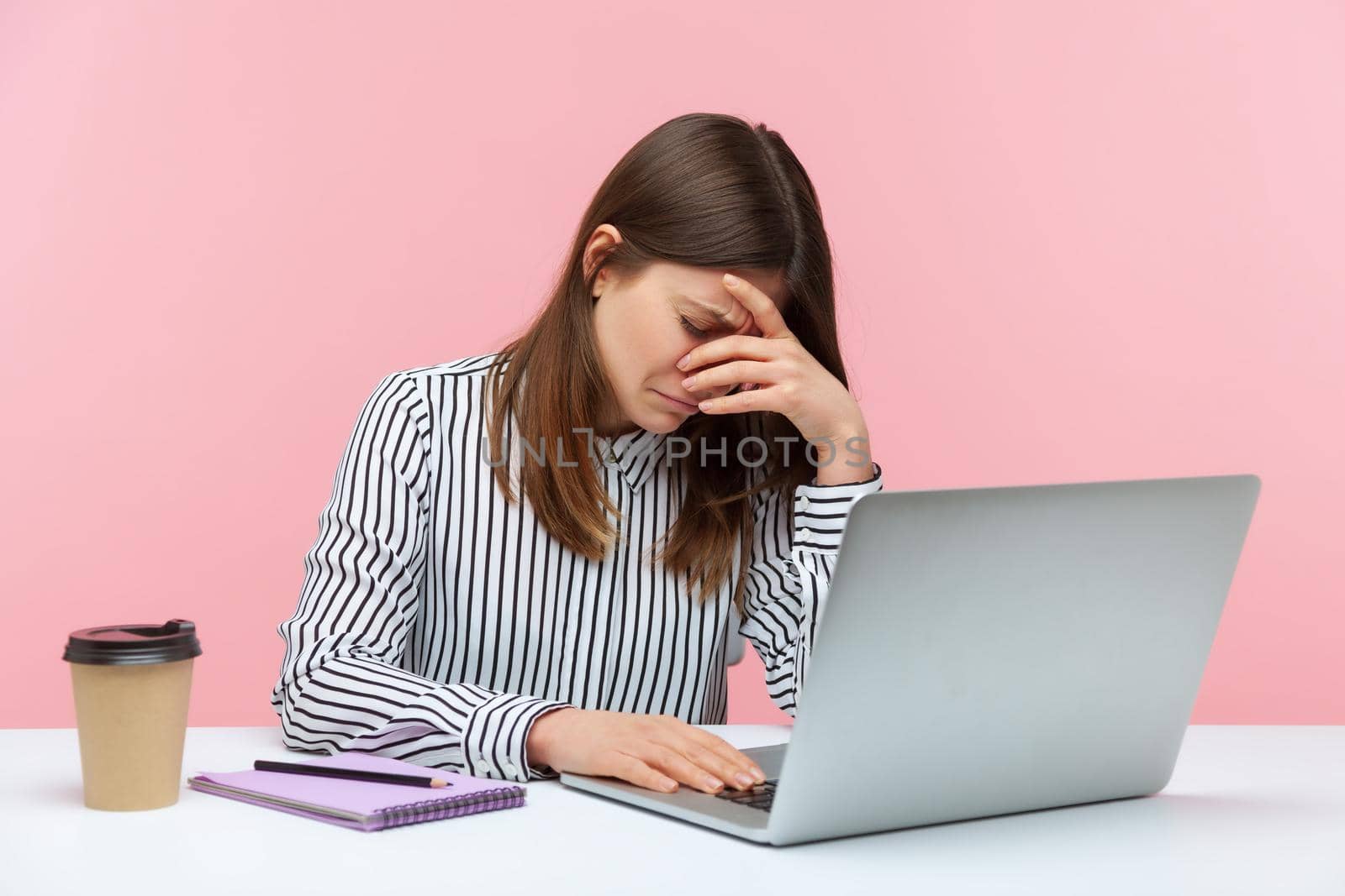 Depressed overworked woman office worker crying leaning head on hand sitting at laptop, feeling worried and desperate about job troubles. Indoor studio shot isolated on pink background