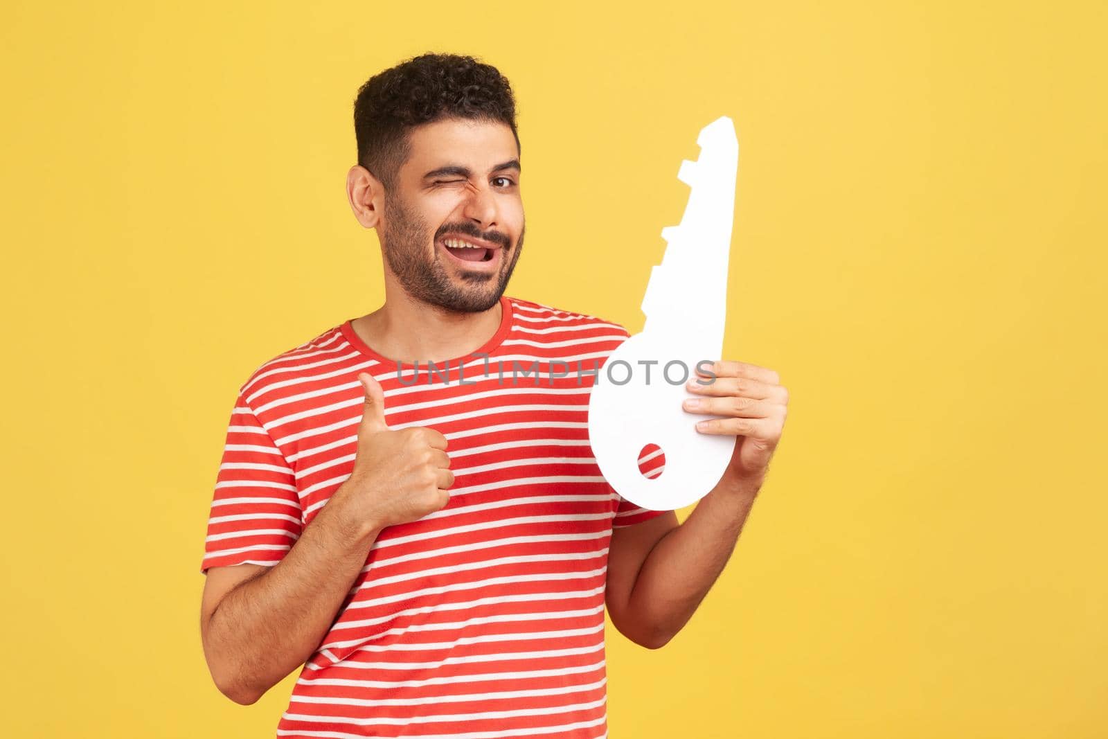 Satisfied bearded man in striped t-shirt showing thumbs up holding huge paper key and smiling, satisfied with new home, real estate purchase, service. Indoor studio shot isolated on yellow background