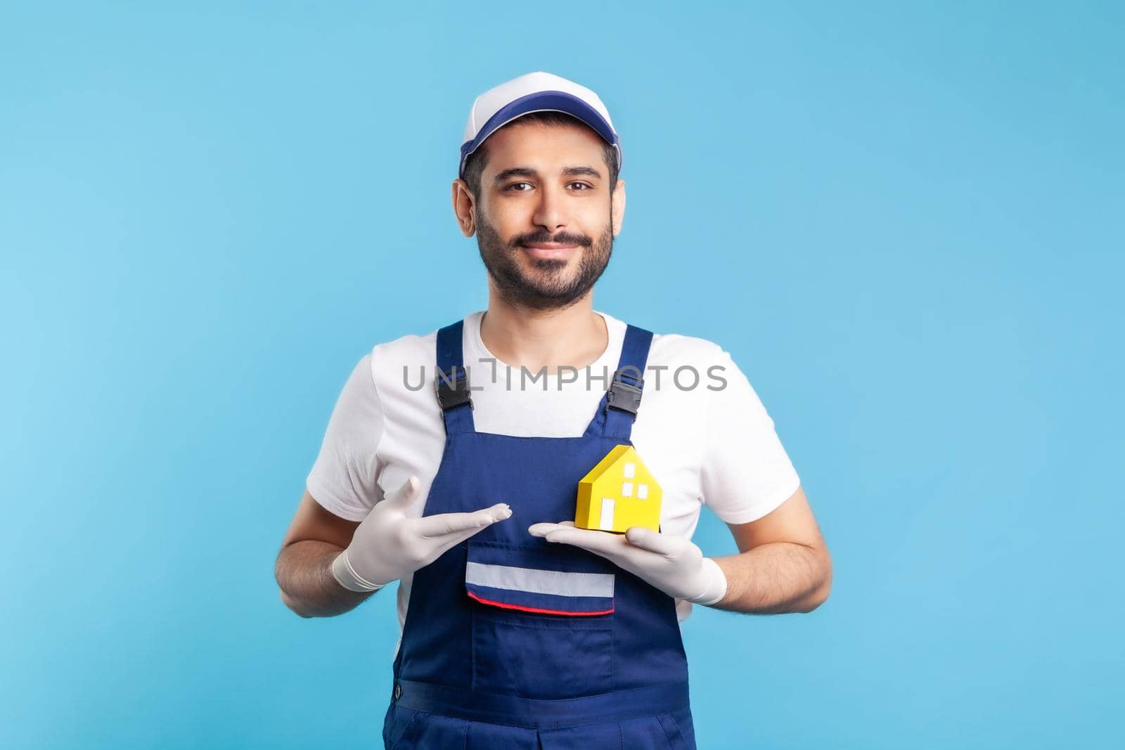 Portrait of happy handyman, mover guy in workwear and gloves showing paper house. Advertising home repair, maintenance or moving services, help in relocation. studio shot isolated on blue background