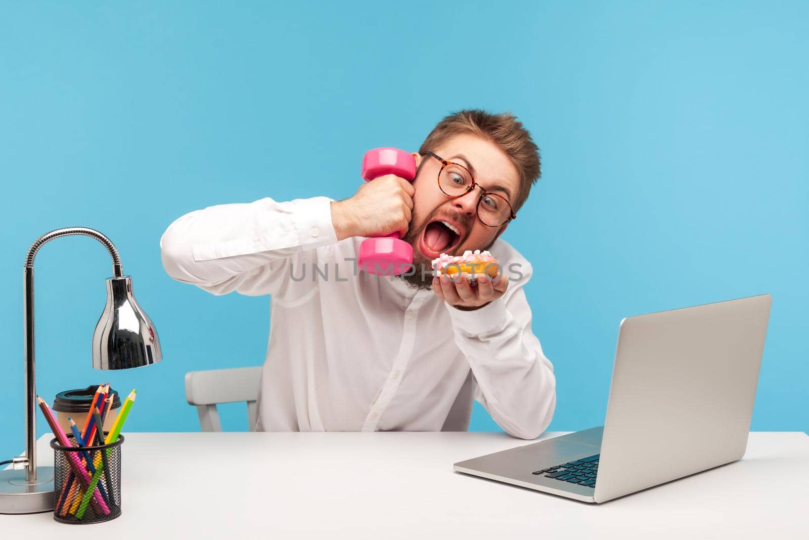 Funny crazy man office worker holding dumbbell but wanting to eat sweet donut ignoring training, temptation, unhealthy eating during work. Indoor studio shot isolated on blue background