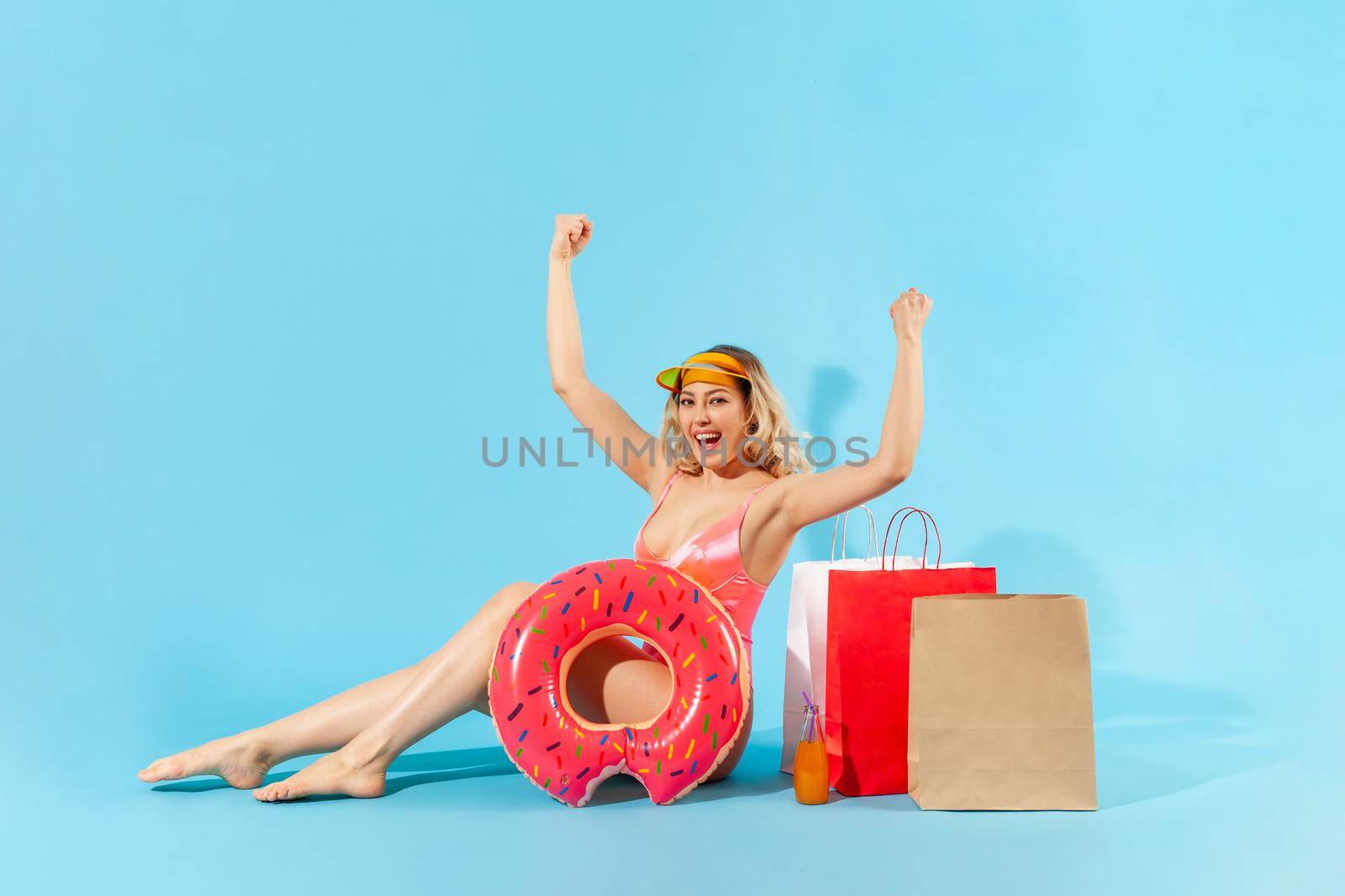 Portrait of enthusiastic overjoyed girl in swimsuit sitting with rubber ring and shopping bags, screaming from happiness, excited by summer sales. indoor studio shot isolated on blue background