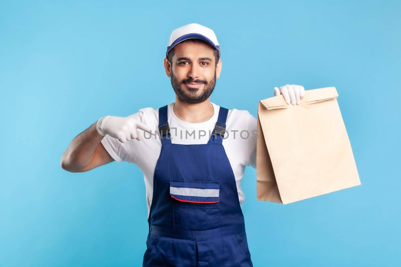 Look at bag. Portrait of happy smiling handyman in overalls and gloves pointing at ordered parcel. Courier delivering food in paper package, post mail services. studio shot isolated on blue background