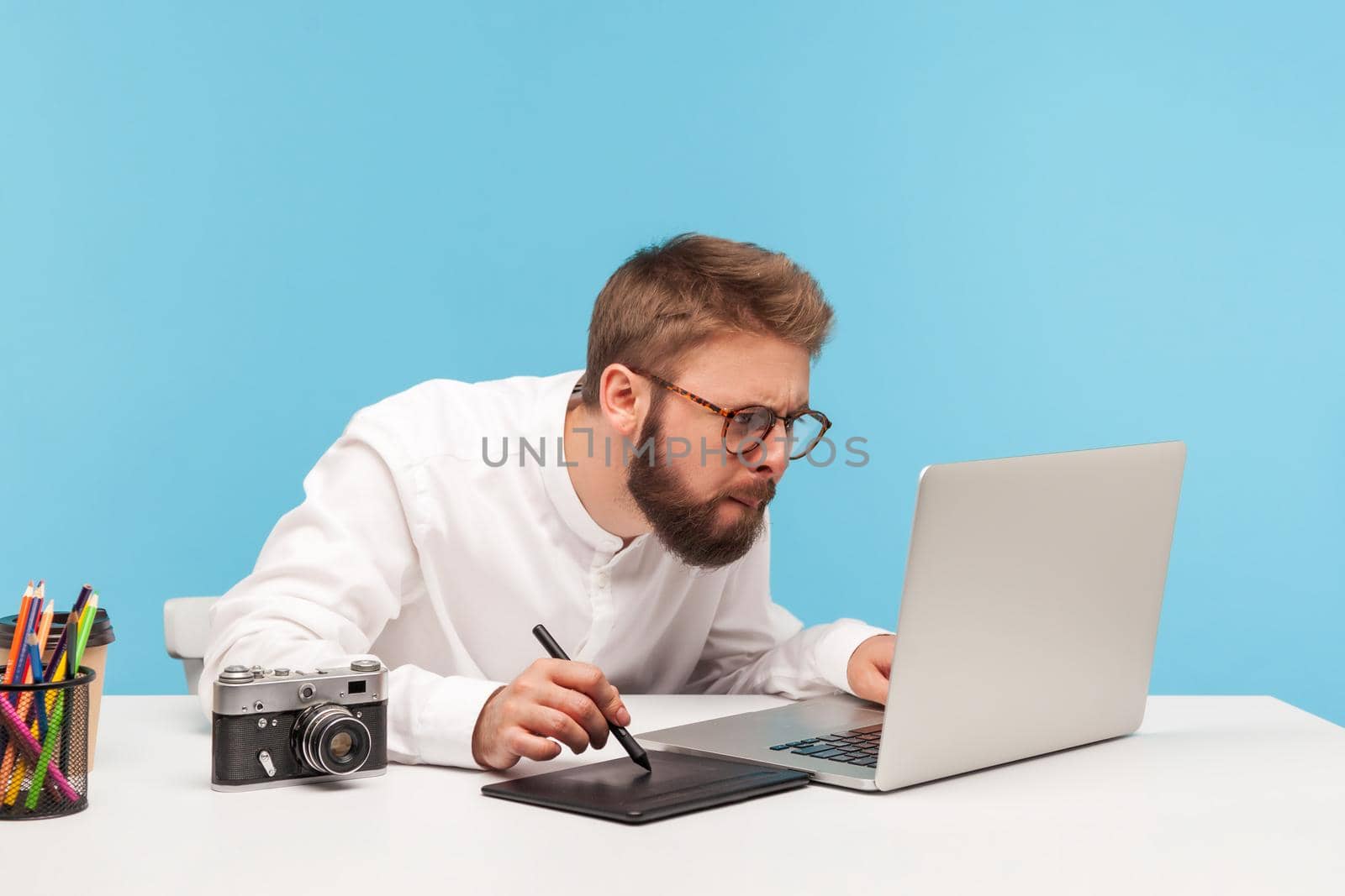 Concentrated bearded man designer drawing on professional tablet looking at laptop display, creating project sitting at workplace. Indoor studio shot isolated on blue background