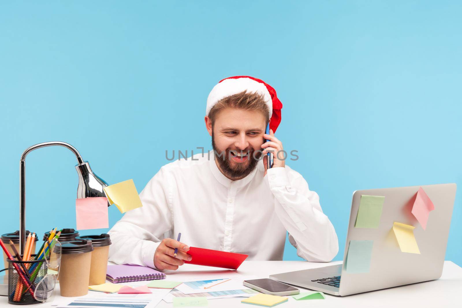 Smiling positive man with beard in santa claus hat talking smartphone holding in hand red envelope with letter and post card, sitting at workplace. Indoor studio shot isolated on blue background