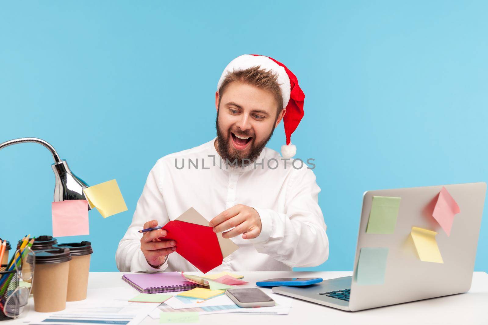 Excited bearded man in santa claus hat opening red envelope, going to read holiday greeting postcard sitting at workplace with laptop. Indoor studio shot isolated on blue background