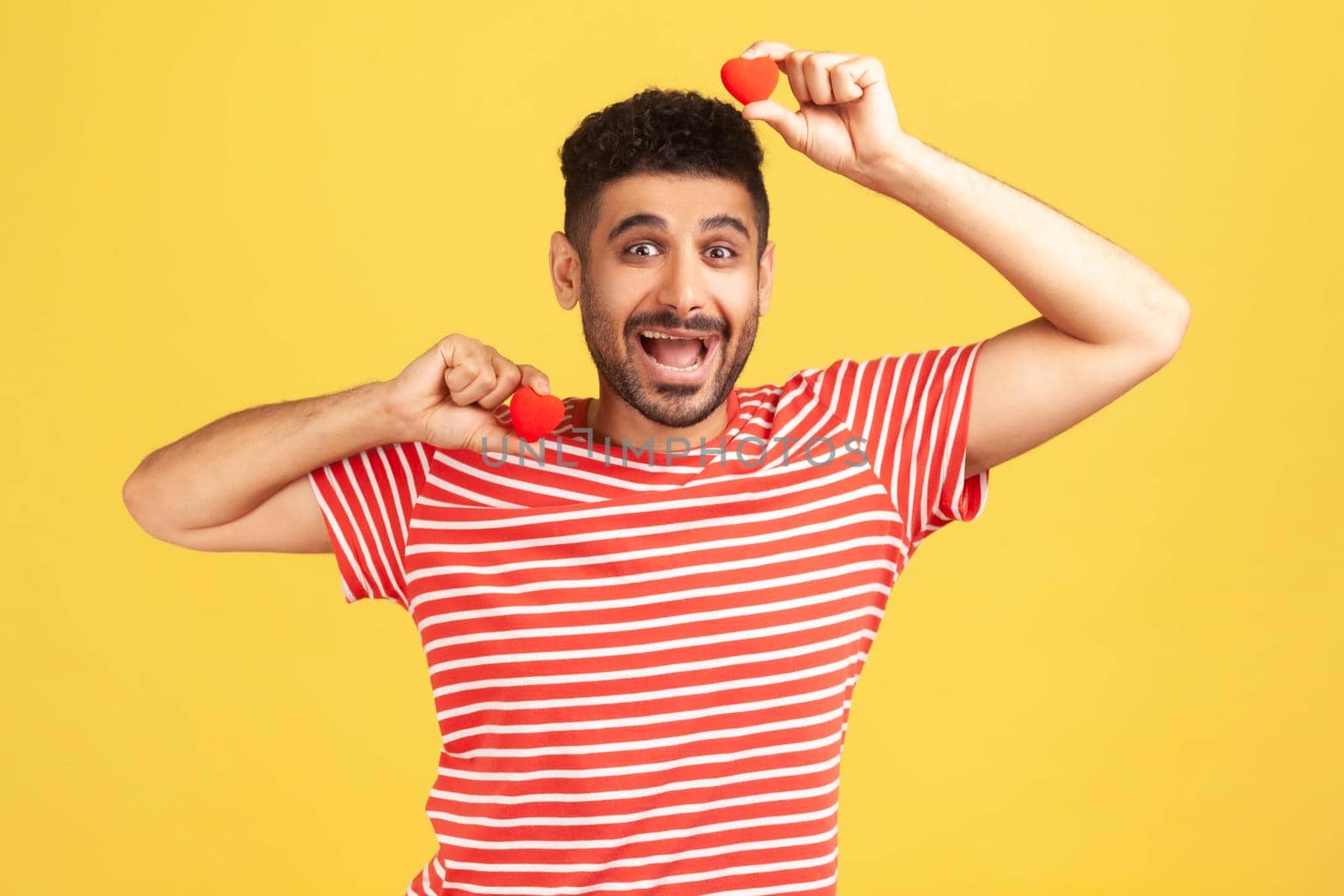 Positive excited bearded man in striped t-shirt fooling around having fun with red toy hearts, showing his fondness and devotion, romance. Indoor studio shot isolated on yellow background