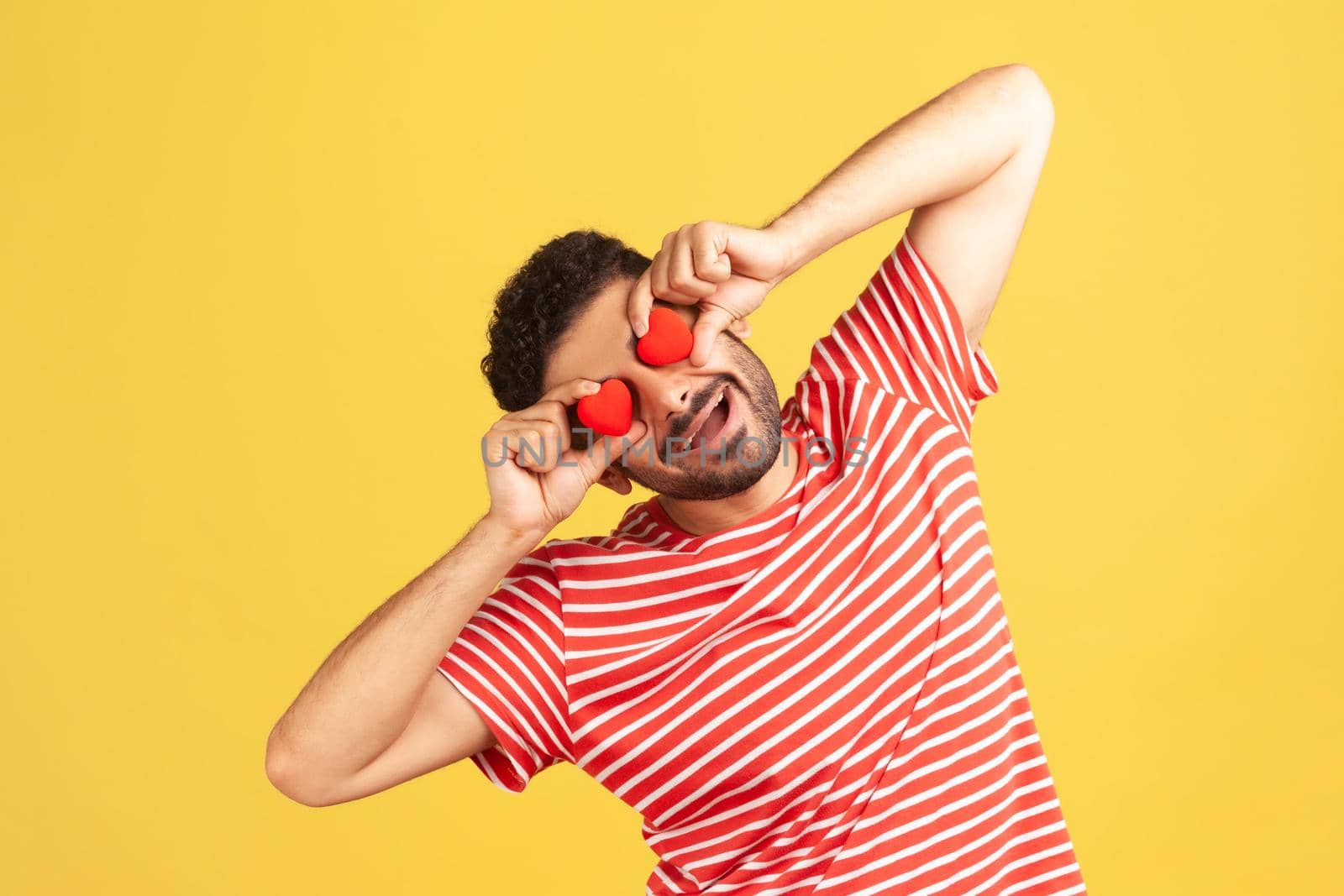 Funny positive man with beard in striped t-shirt having fun holding little toy hearts near eyes and smiling, showing his affection, love confession. Indoor studio shot isolated on yellow background