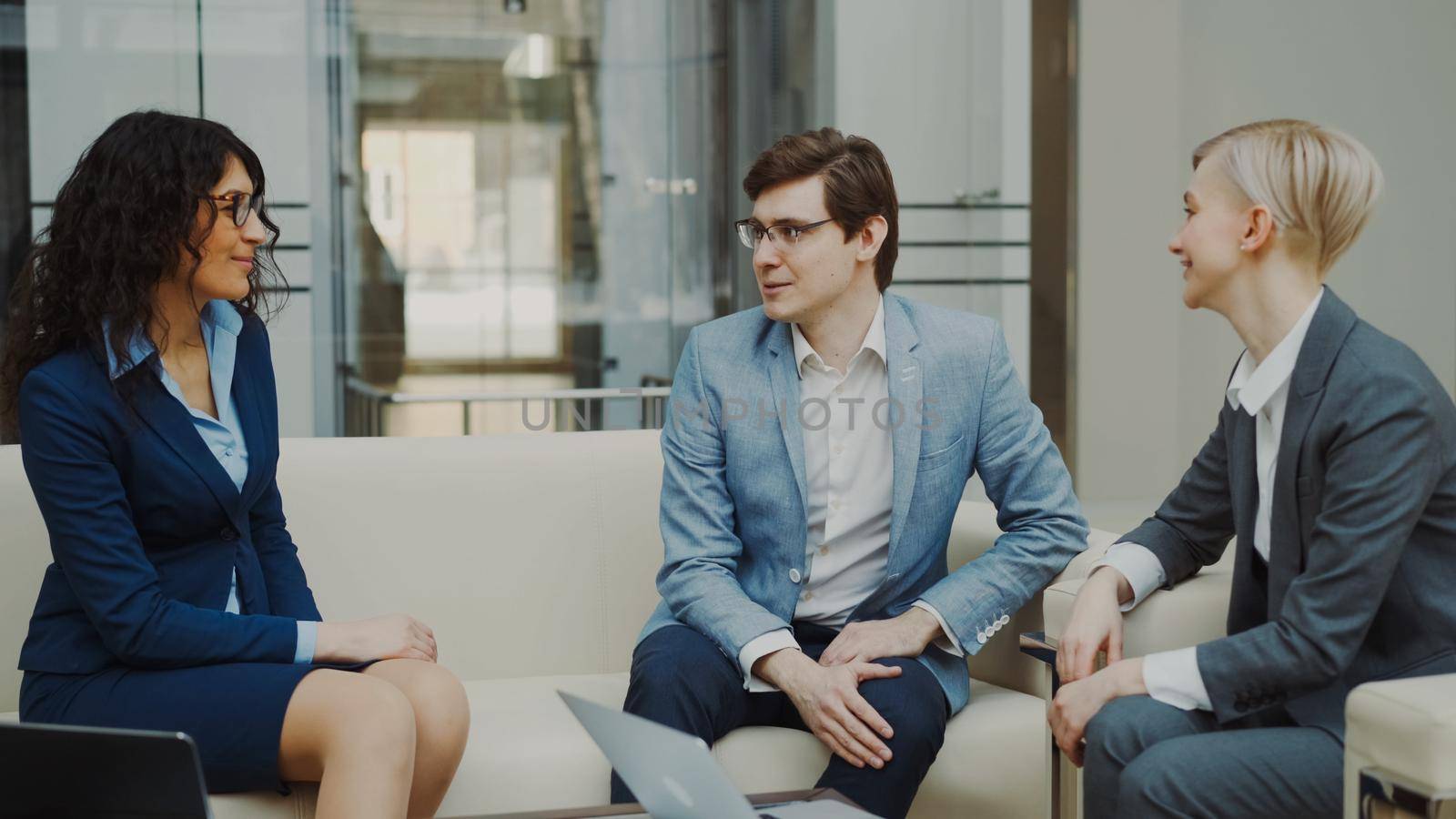 Businessman talking and duscussing future contract with female business partners sitting on couch in meeting room by silverkblack