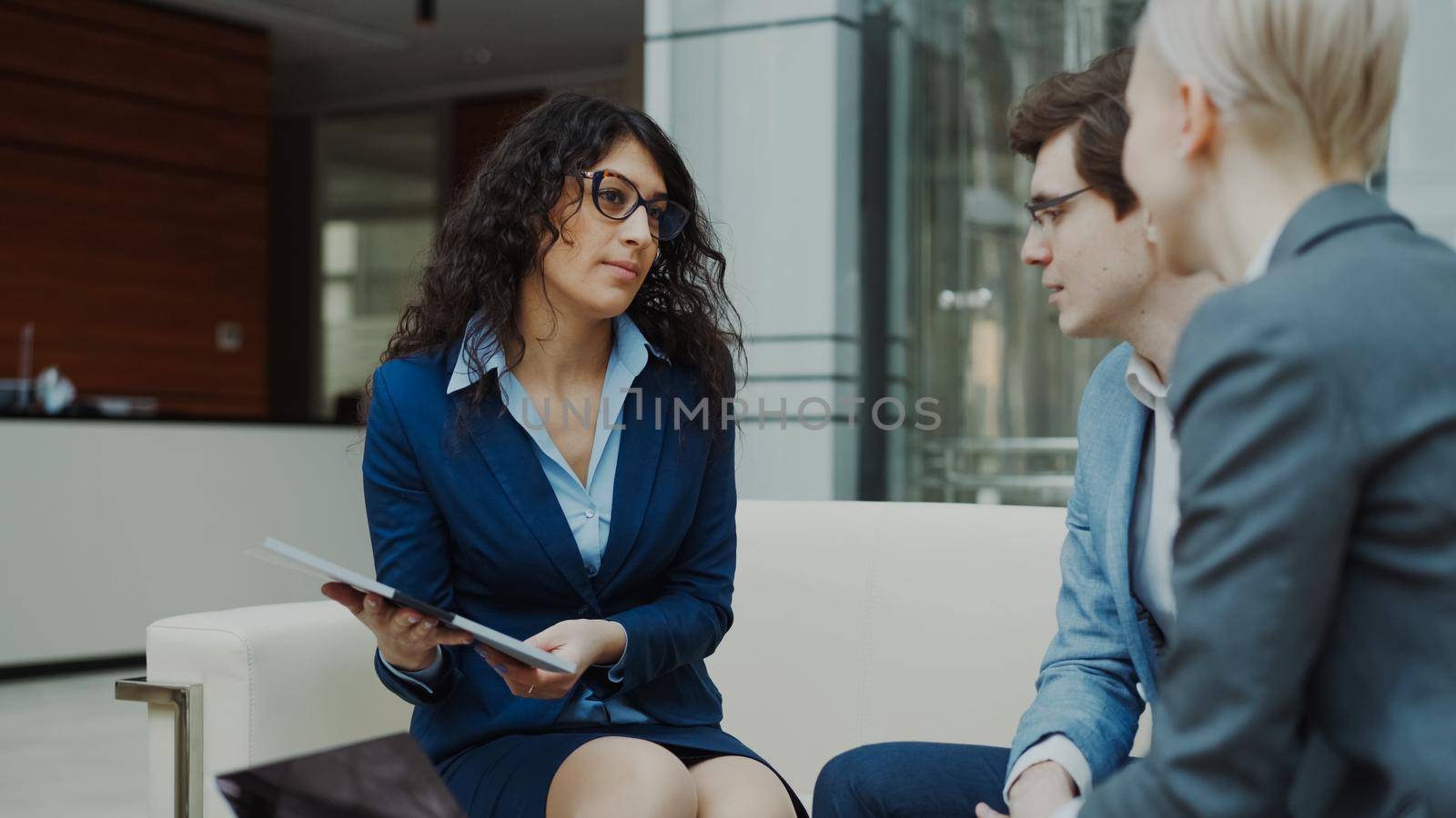 Businesswoman in glasses talking and duscussing future contract with business partners sitting on couch in modern office indoors