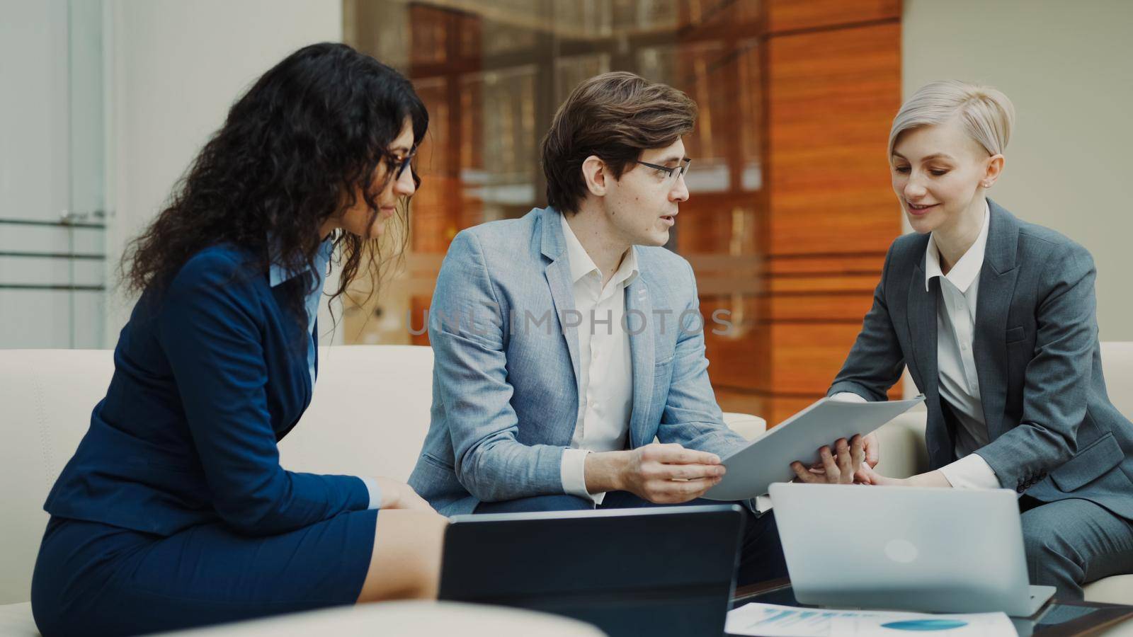 Businessman in glasses talking and duscussing future contract with female business partners sitting on couch in meeting room indoors