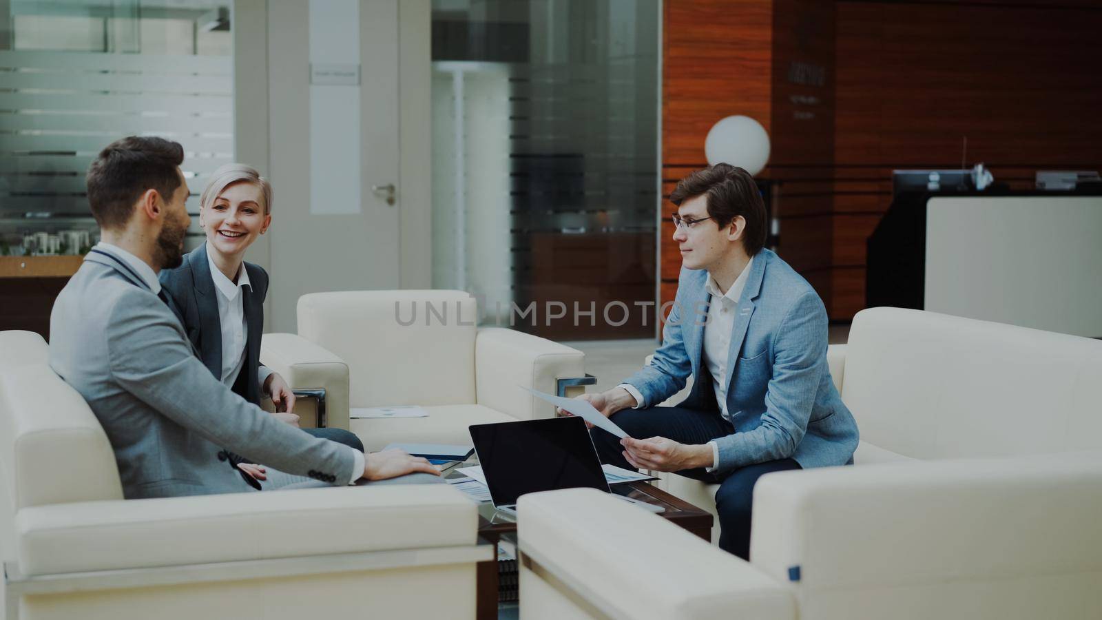 Cheerful businessman discussing financial reports with business partners sitting on sofa in modern office hall by silverkblack