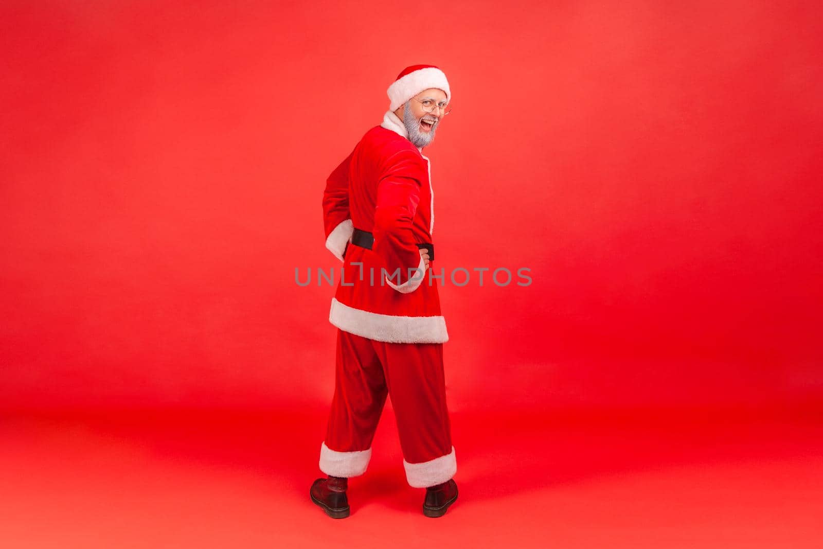 Full length portrait of elderly man with gray beard in santa claus costume standing backwards with hands on hips, turning to camera with excited look. Indoor studio shot isolated on red background.