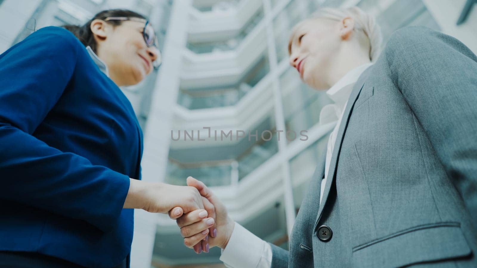 Low angle shot of businesswoman shaking hands and talking with female business colleague in suit in hall of modern office building indoors