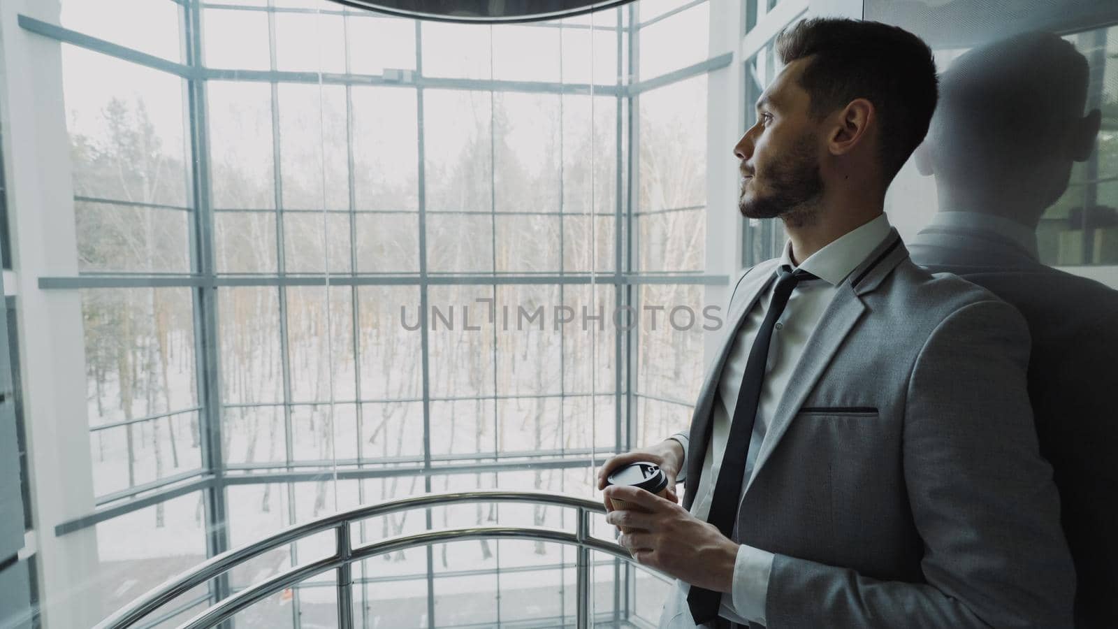 Young handsome businessman in suit drinking coffee moving up inside eleavator in modern business center indoors