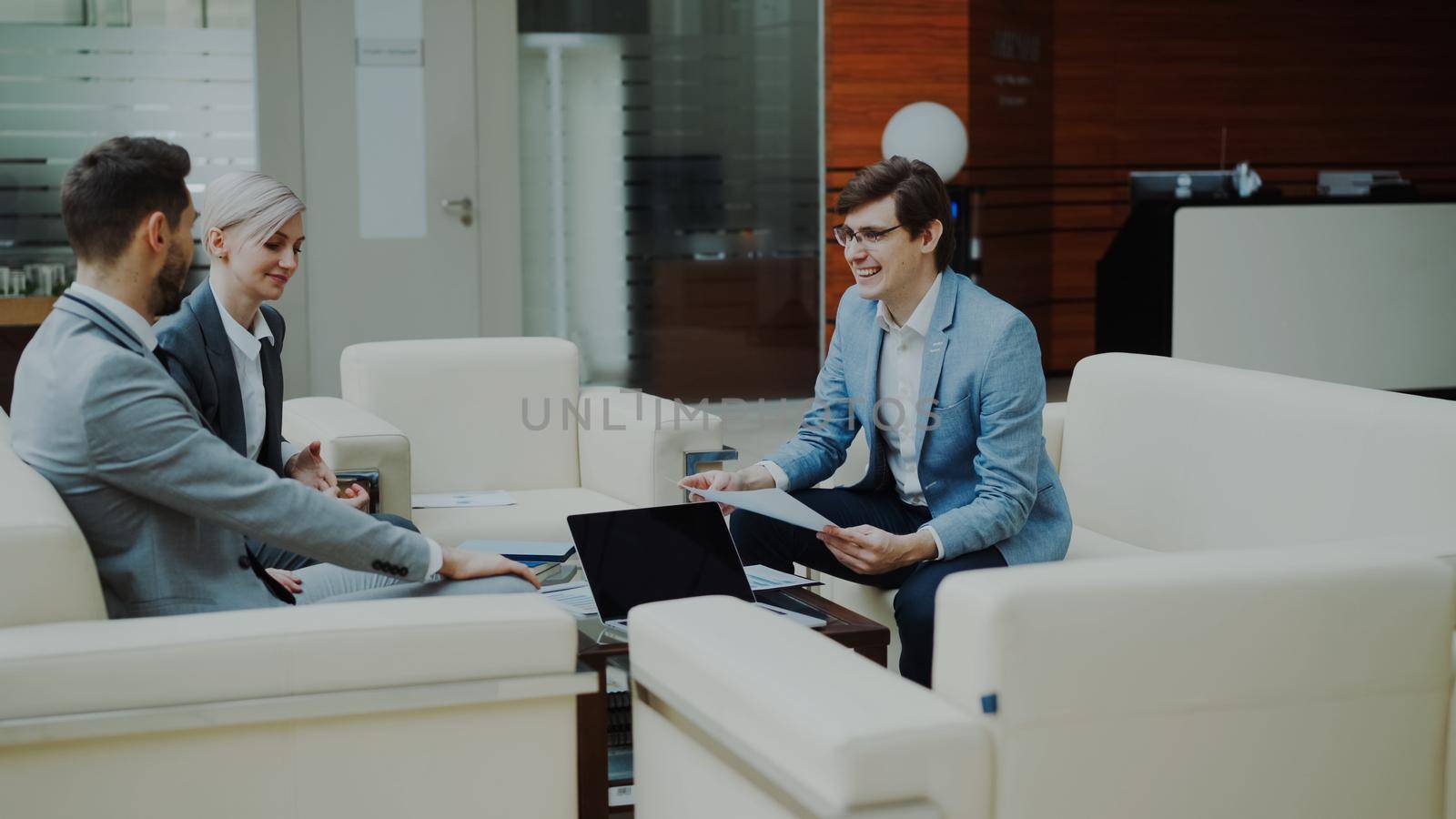 Cheerful businessman discussing financial reports with business partners sitting on sofa in modern office hall indoors