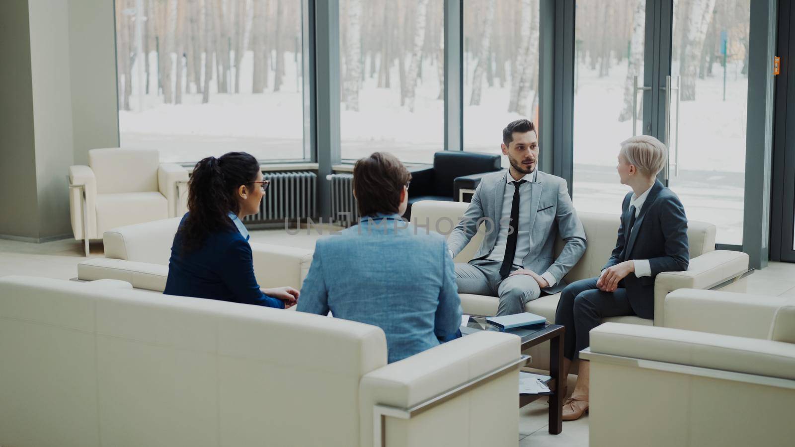 Group of business partners discussing agreement whilesitting on couch in modern office hall with panoramic windows indoors