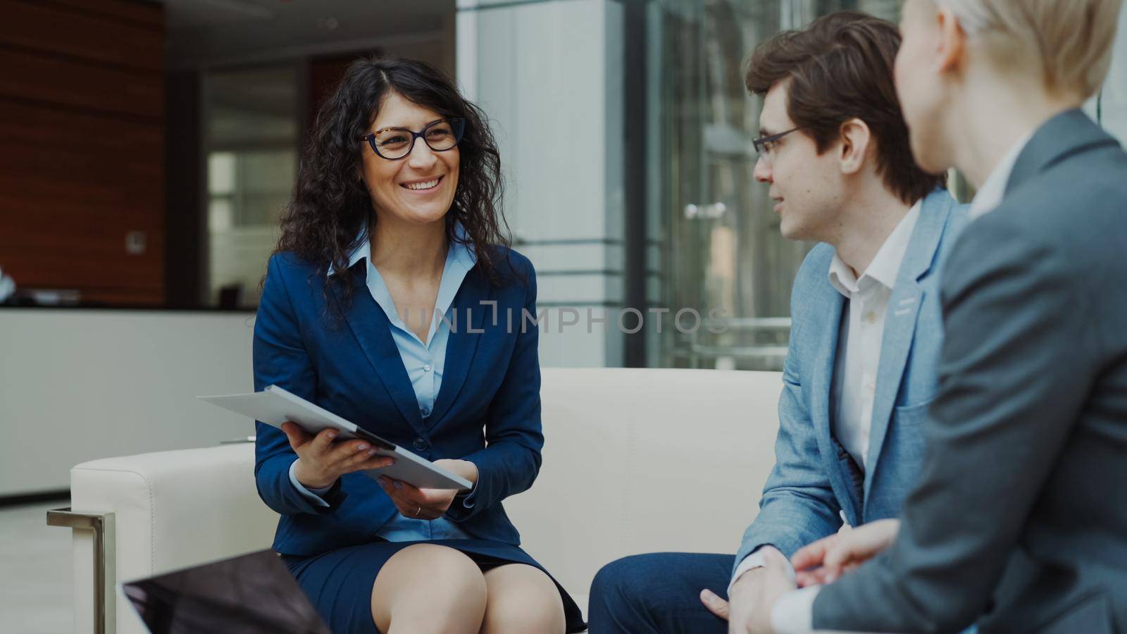 Businesswoman in glasses talking and duscussing future contract with business partners sitting on couch in modern office indoors