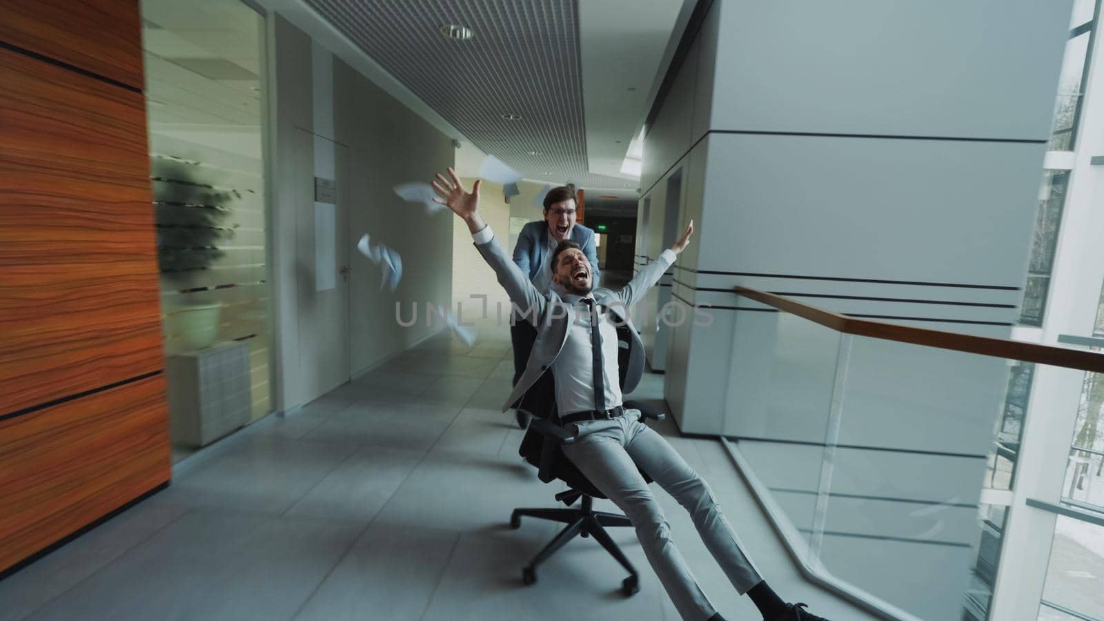 Two crazy businessmen riding office chair and throwing papers up while having fun in lobby of modern business center indoors