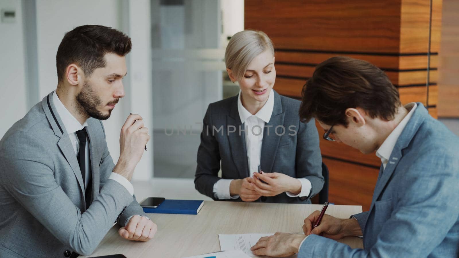 Young businessman in suit signing contract with his colleague while corporate lawyer explaining details of partnership sitting in modern office indoors